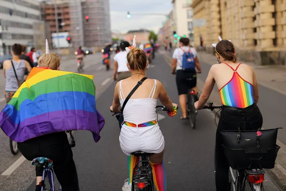 Mehrere junge Menschen fahren gemeinsam auf Fahrrädern eine breite Straße entlang, umgeben von städtischen Gebäuden. Sie tragen Kleidung mit Regenbogenfarben. Eine Person ist in eine Regenbogenflagge gehüllt.