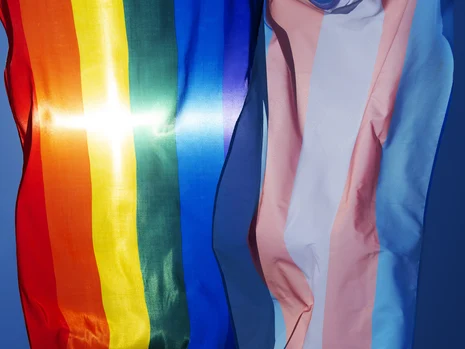 Zwei Fahnen, Regenbogenflagge und Trans-Flagge, wehen vor blauem Himmel. Sonnenlicht spiegelt auf dem Stoff. Die Aufnahme vermittelt Sichtbarkeit, Vielfalt und Stolz queerer und trans Communitys.