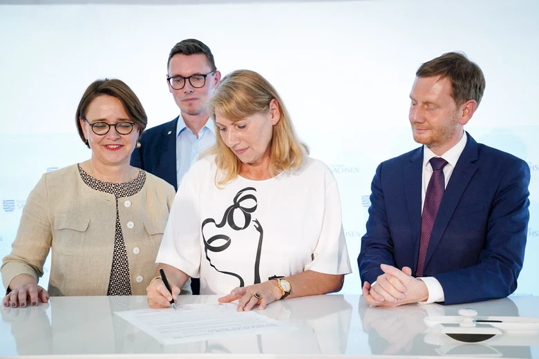 Drei Personen bei einer offiziellen Unterzeichnung: Ein Mann sitzt und unterschreibt, zwei Frauen stehen daneben. Das Setting wirkt freundlich-offiziell und findet in einem hellen, neutralen Raum statt.