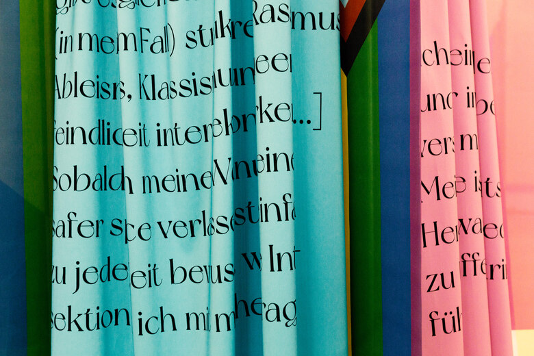 Bunte Stoffbahnen mit großformatiger, schwarzer Textaufschrift hängen nebeneinander. Die Farben erinnern an Pride-Flaggen und thematisieren Vielfalt und Zugehörigkeit auf künstlerische Weise.