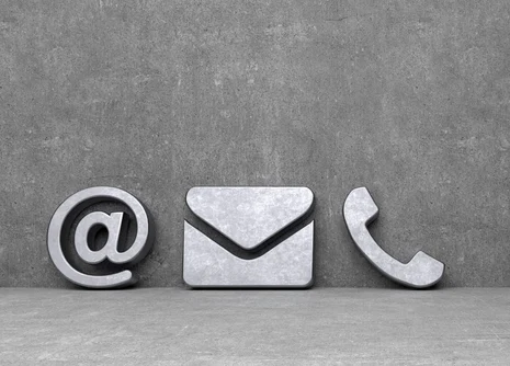 Drei graue Symbole vor Betonwand: ein @-Zeichen, ein Briefumschlag und ein Telefonhörer. Sie stehen nebeneinander und symbolisieren moderne Kontaktmöglichkeiten per E-Mail und Telefon.